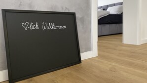 Une pancarte d’accueil à cadre noir, sur laquelle est écrit le mot « Willkommen », est placée dans une pièce où se trouvent un canapé gris et un plancher en bois.