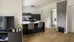 Une cuisine moderne avec un îlot central, des tabourets de bar et une table à manger. On y trouve un téléviseur posé sur un meuble et une plante sur le comptoir.