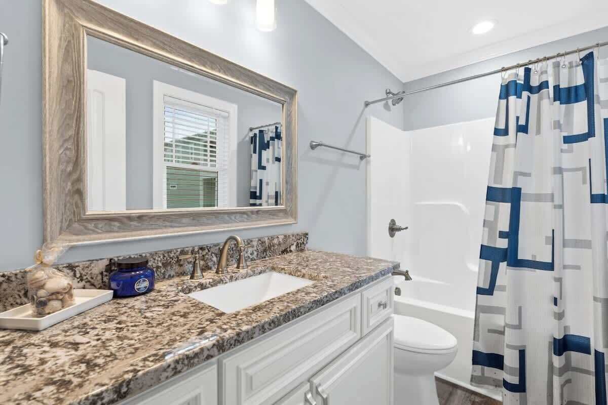 Une salle de bain avec un lavabo surmonté d’un plan de travail en granit, des toilettes blanches, une douche avec un rideau à motifs bleus et blancs, et un grand miroir avec un cadre en bois.