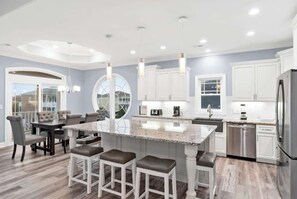 A modern kitchen with a central island, bar stools, and a dining area with a round table and chairs. - East End Views (Ocean Isle Beach)