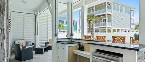 A covered outdoor kitchen with a sink, a dining area with chairs, and a view of a building with balconies.