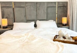 A neatly made bed with white linens, a tray with a teapot, cups, and a pastry, and two bedside lamps on a modern room with a grey headboard.