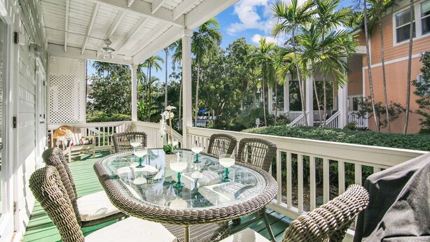Une terrasse extérieure couverte, aménagée avec des meubles en osier, une table en verre et offrant une vue sur des palmiers et un bâtiment.