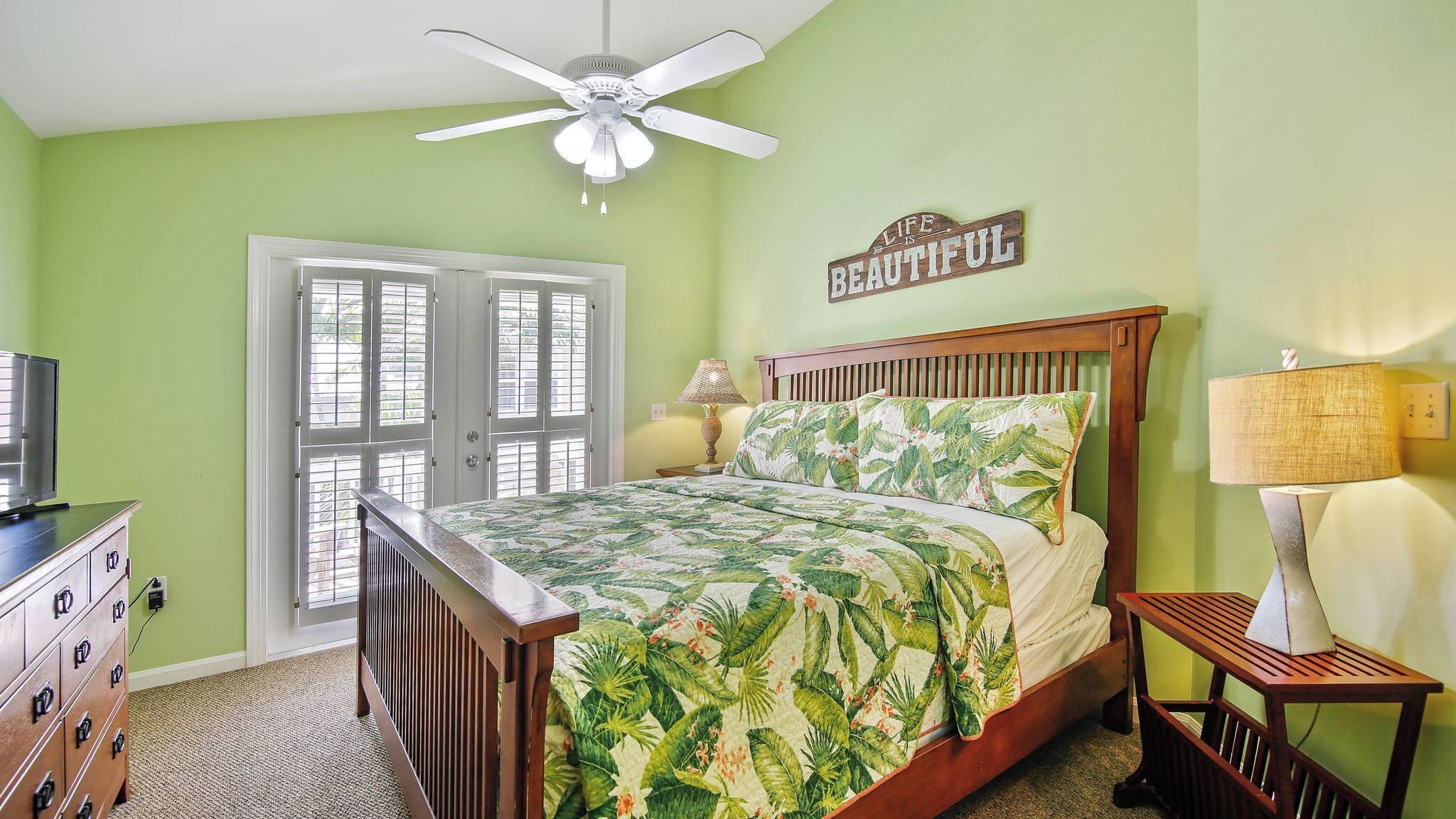 A bedroom with a bed, a ceiling fan, a nightstand with a lamp, a dresser, and a door with windows.