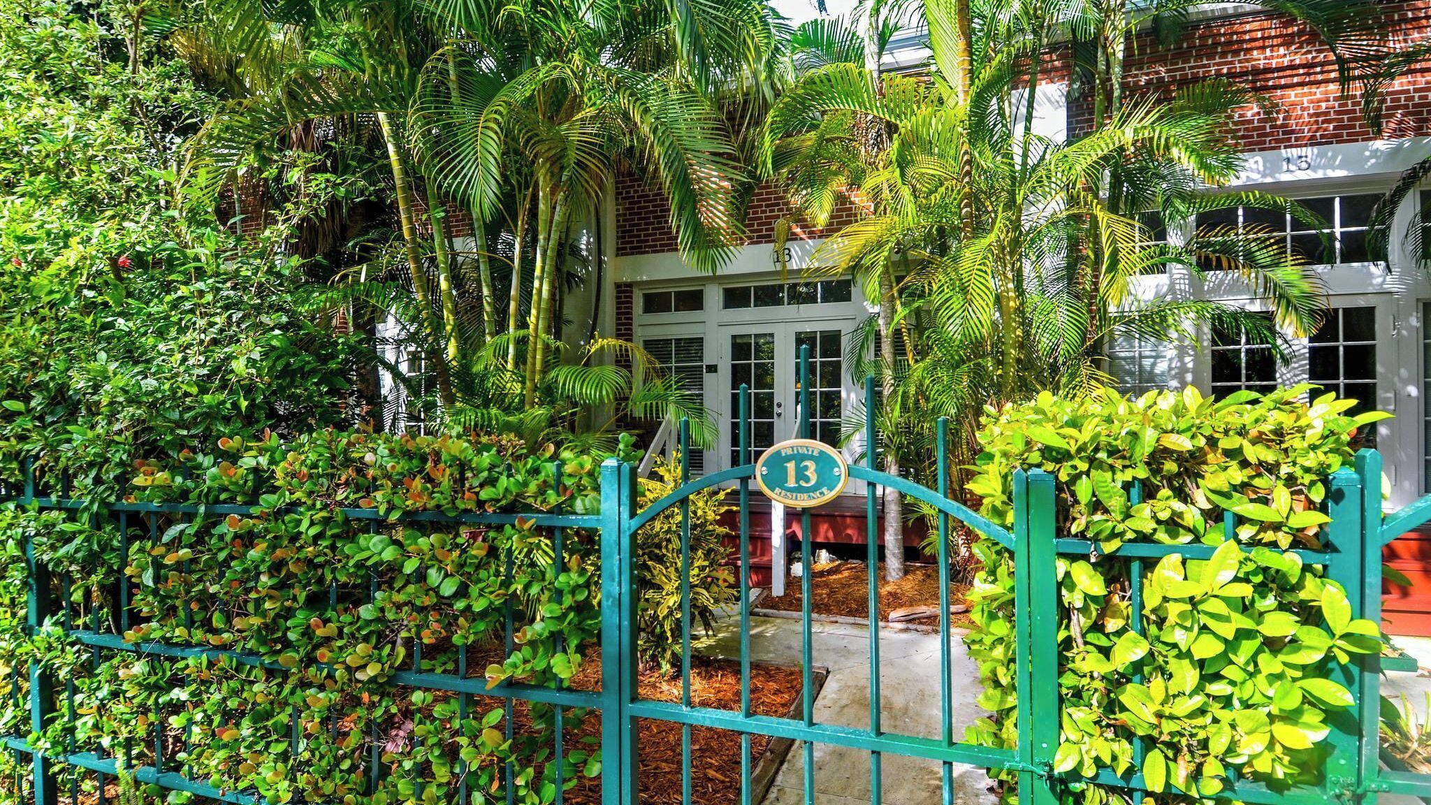 A private residence with a green metal fence, lush tropical plants, and a brick building in the background.