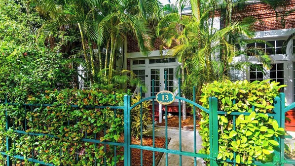 A private residence with a green metal fence, lush greenery, and a brick building in the background. - Key West Found by Avantstay Close to Shops w/ Patio & Shared Pool! Month Long Stays Only (Key West)
