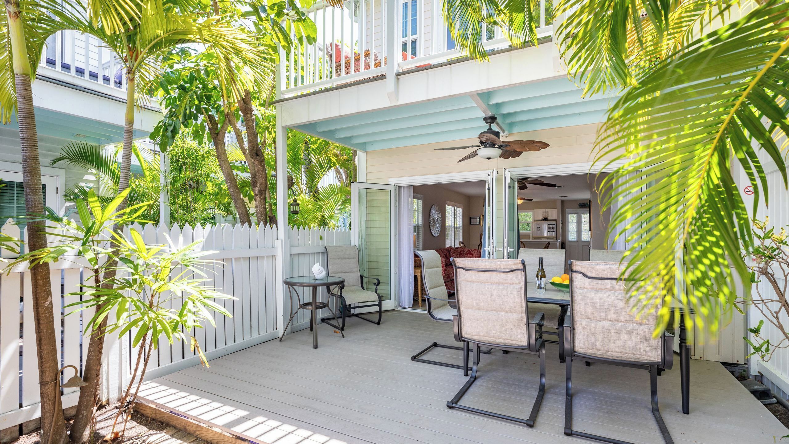 A covered outdoor patio with a dining table and chairs, surrounded by palm trees and a white picket fence.