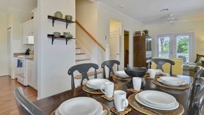 Un coin repas avec une table en bois dressée pour quatre personnes, entourée de chaises. On aperçoit un escalier au fond, qui mène à un autre niveau.