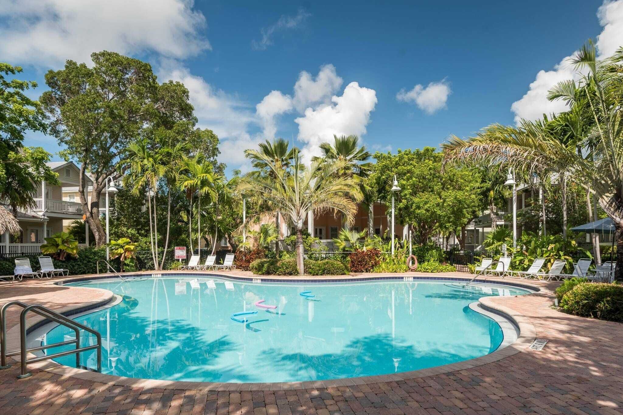 A swimming pool surrounded by palm trees and lounge chairs, with a clear blue sky above.