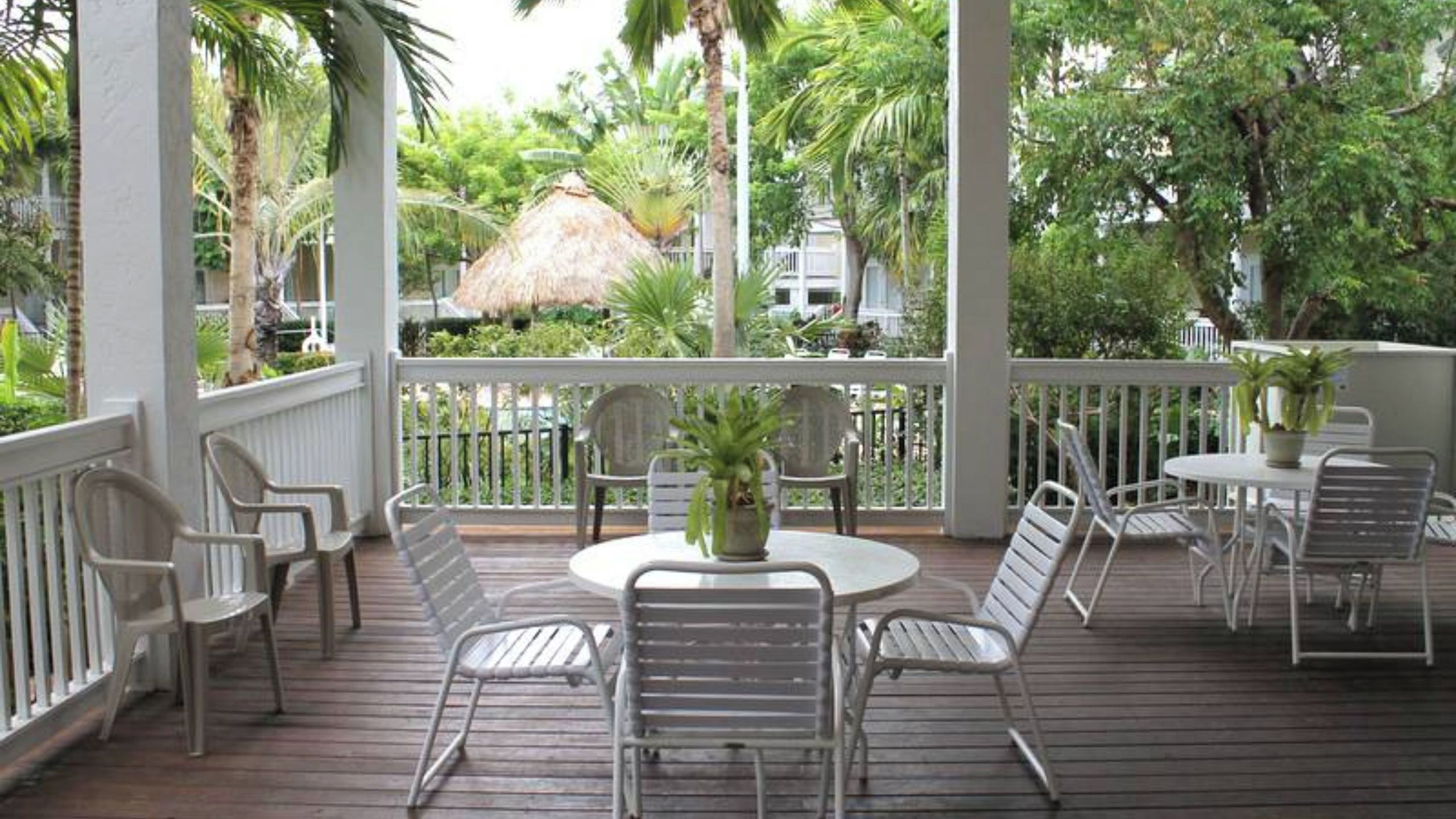 A covered outdoor area with a wooden deck, white metal chairs, a round table, and potted plants.