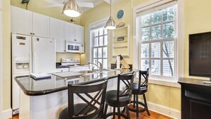 A kitchen with white cabinets, a microwave, a stove, a sink, and a bar area with stools. There are two pendant lights, a clock, and a window with a view of trees. - Lazy Gecko by Avantstay Walk to Duval St in Key West! Month Long Stays Only (Key West)