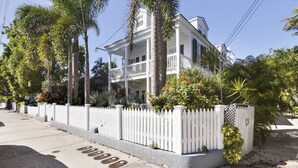 A two-story white house with a balcony, surrounded by a white picket fence and palm trees. - Lazy Gecko by Avantstay Walk to Duval St in Key West! Month Long Stays Only (Key West)