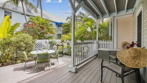 Een overdekt terras met een witte balustrade, groene stoelen en een tafel met een rieten zitting.