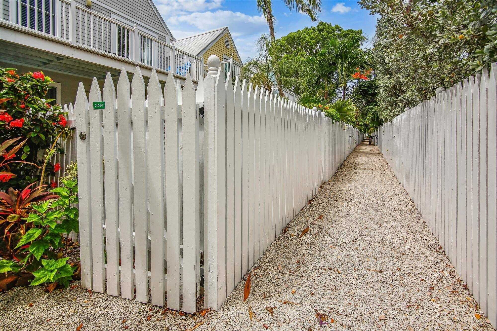 A narrow gravel pathway flanked by white picket fences leading to a building with a balcony.