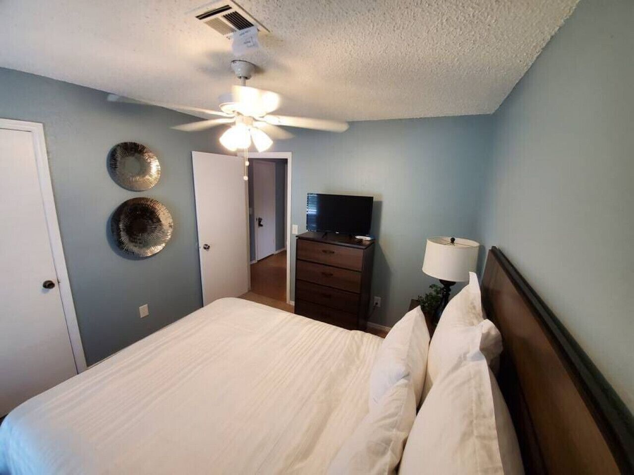 Ein Schlafzimmer mit einem Bett, einem Deckenventilator, einem Schminktisch, einem Fernseher und einer Tür.