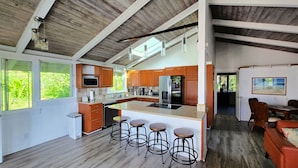 A modern kitchen with wooden cabinets, a central island with stools, and a dining area with a view of greenery. - Quiet 3-bedroom house on 2.5 acres w/ocean and town views, 6 beds (Kailua-Kona)