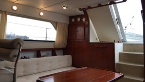 Camarote de barco con un banco acolchado, una mesa de madera, una ventana con cortinas y una puerta con espejo.