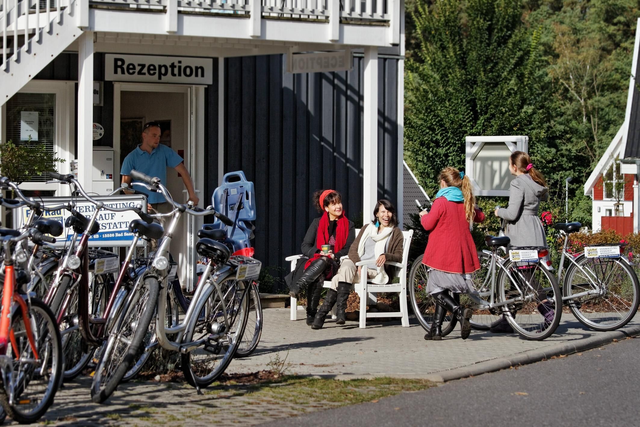 Un edificio con un cartel que dice 'Rezeption', bicicletas estacionadas afuera y un grupo de personas sentadas en bancos.