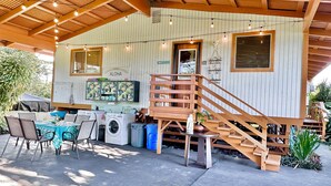 Un espace extérieur abrité, doté d’un escalier en bois, d’une table avec des chaises, d’un lave-linge et d’une enseigne indiquant « Hawaii Aloha ».