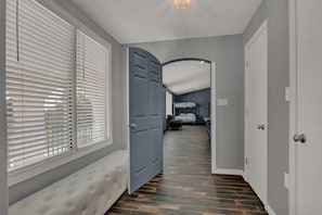 A hallway with a bench, a door leading to a bedroom, and a view of the outside through blinds.