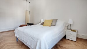 A modern bedroom with a large bed, white bedding, a nightstand with a lamp, and a wooden floor. - Am Pavillon Bed & Kitchen (Bern)
