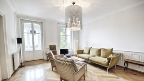 A spacious living room with a herringbone wood floor, a beige sofa, two striped armchairs, a coffee table, and a large chandelier. - Am Pavillon Bed & Kitchen (Bern)
