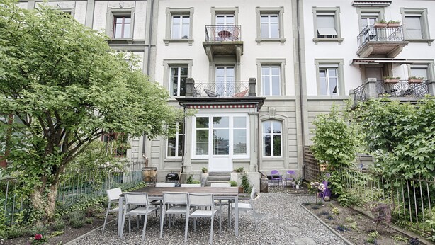 A multi-story building with a garden patio area featuring a table and chairs, surrounded by a gravel path and various plants. - Am Pavillon Bed & Kitchen (Bern)