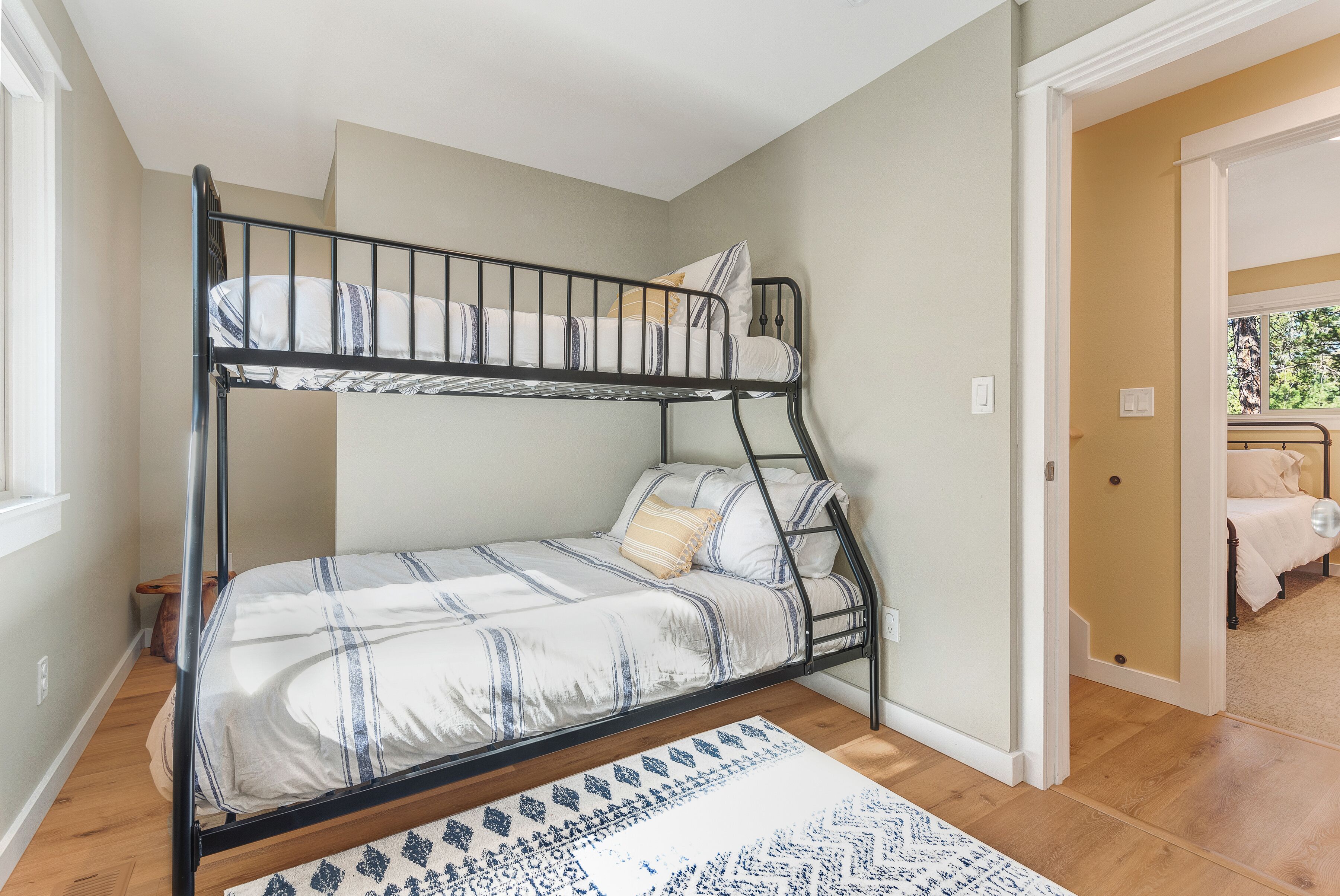 Een stapelbed met een gebedrukte sprei en kussens, een houten vloer, en een uitzicht op een gang die naar een andere kamer leidt.