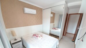 A hotel room with a bed, a nightstand, a lamp, a mirror, and a door leading to another room. - 314, Excelente Apartamento Para Desfrutar Férias (Bombinhas)