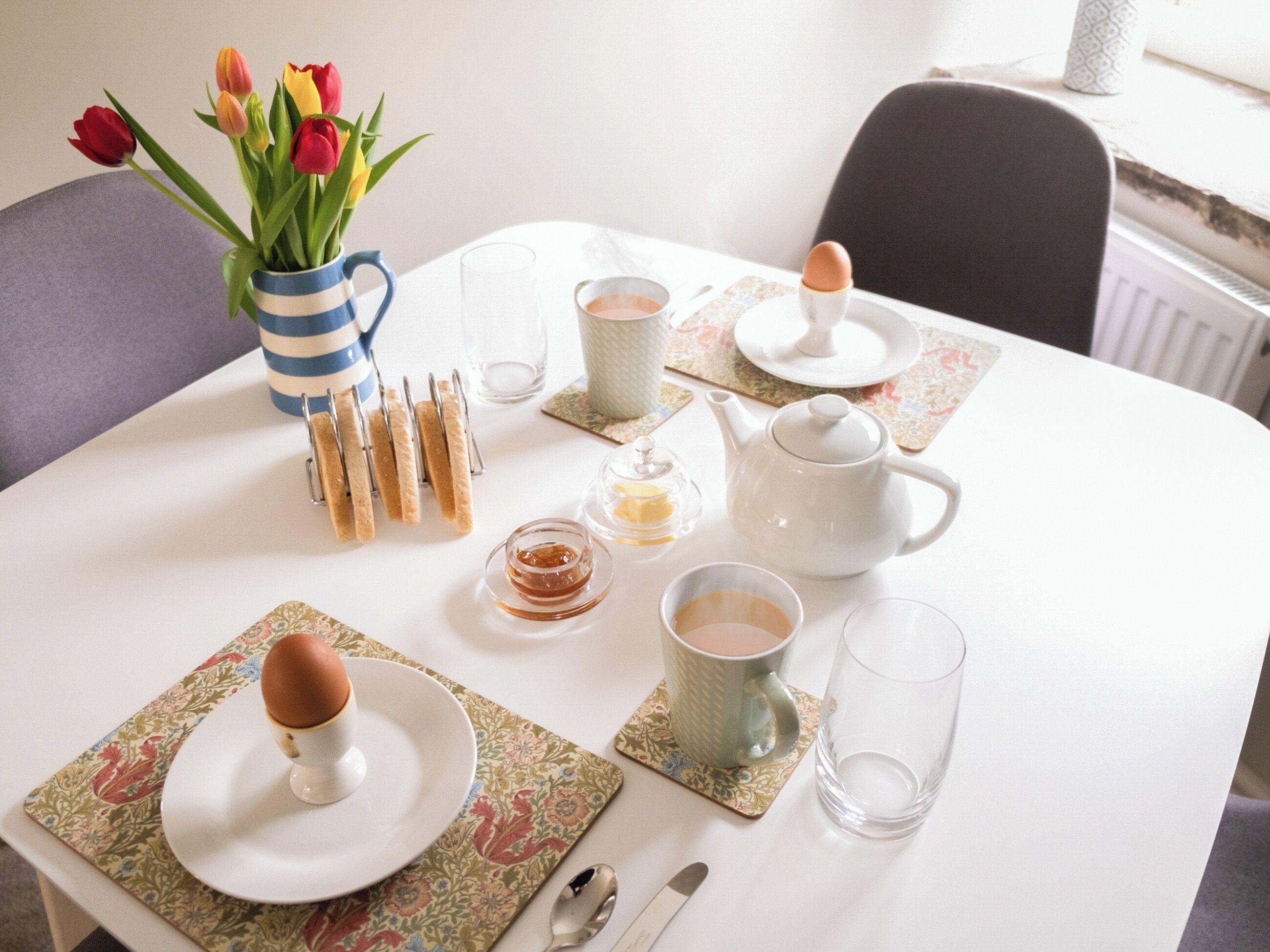 Ein Esstisch ist für das Frühstück gedeckt, mit einer Teekanne, Tassen, einem Teller mit einem Ei und einer Vase mit Tulpen.