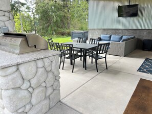 An outdoor patio with a stone wall, a grill, a table with chairs, a sofa, and a TV mounted on a corrugated metal wall. - Water View Modern Retreat- Hot Tub + Scenic Views (Gig Harbor)