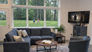 Uma sala de estar com um sofá modular cinza escuro, uma mesa de centro redonda e uma poltrona preta. Uma televisão está montada na parede e uma estante de livros é visível ao fundo.