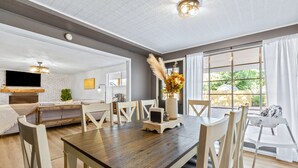 Un comedor moderno con una mesa de madera y sillas blancas, un televisor montado en una pared de ladrillo blanco, y un ventanal con cortinas.