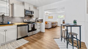 Una cocina moderna con armarios blancos, electrodomésticos de acero inoxidable y encimera de madera. Hay una zona de comedor con una mesa y taburetes de bar.
