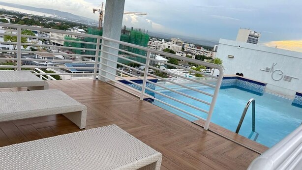 Un área de piscina en la azotea con sillones blancos, piscina con azulejos azules y vista panorámica de la ciudad.