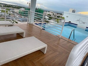 Un área de piscina en la azotea con sillones blancos, piscina con azulejos azules y vista panorámica de la ciudad.
