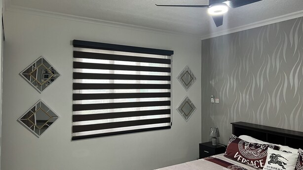 Un dormitorio con un ventanal grande cubierto por persianas horizontales, un ventilador de techo, una cama con cabecero y espejos decorativos en la pared.
