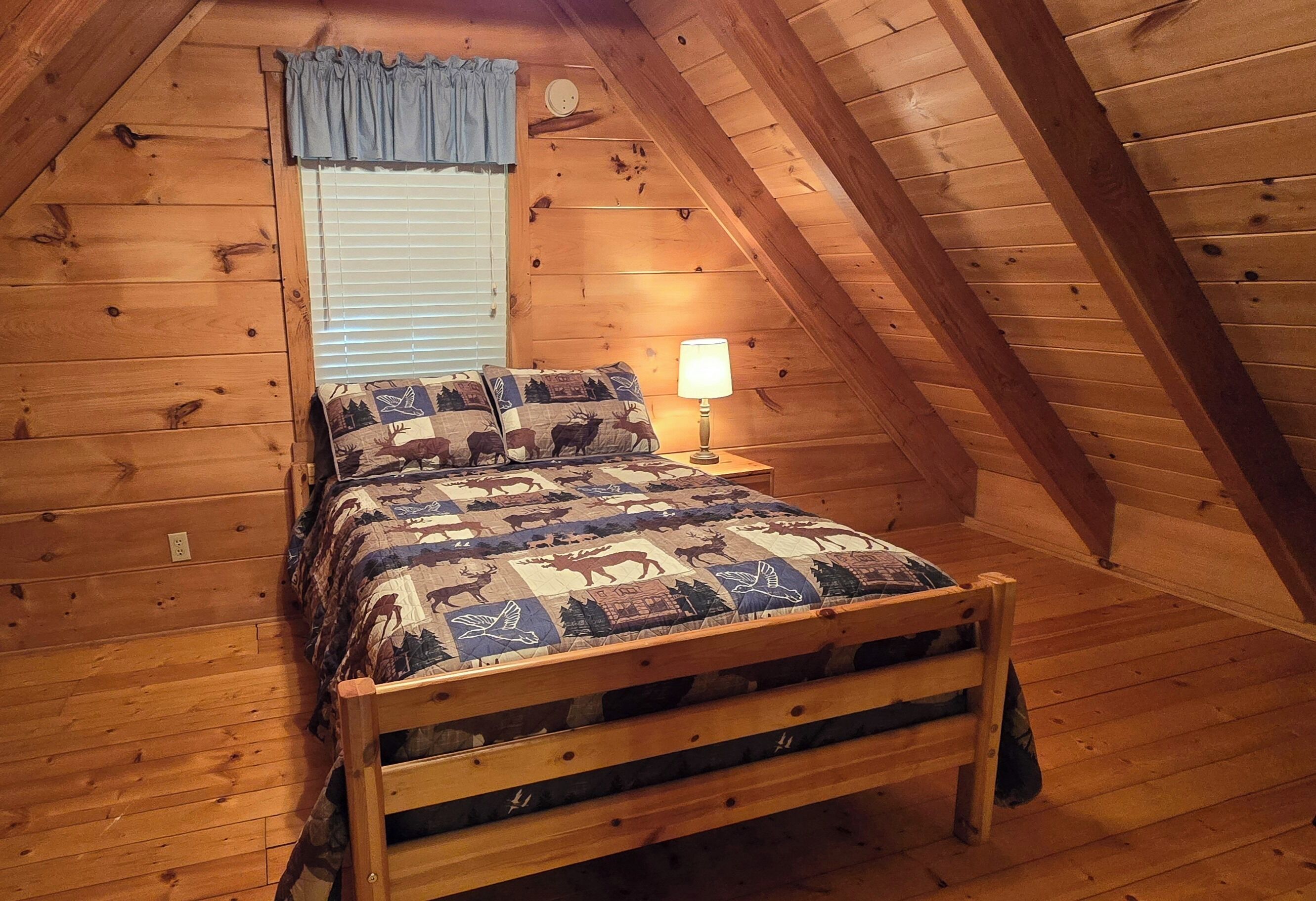 A cozy attic bedroom with wooden walls, a bed with a patterned quilt, a bedside lamp, and a window with blinds.