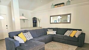 Un salon moderne avec un canapé d’angle gris, un tapis à motifs et un panneau « Bienvenue » accroché au mur.