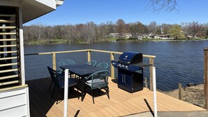 Eine Terrasse mit Grill und Stühlen mit Blick auf einen See, umgeben von Bäumen und Häusern.
