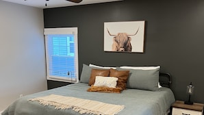 Ein Schlafzimmer mit einem Bett, einem Nachttisch, einem Fenster mit Jalousien und einem Wandbild eines Stiers.