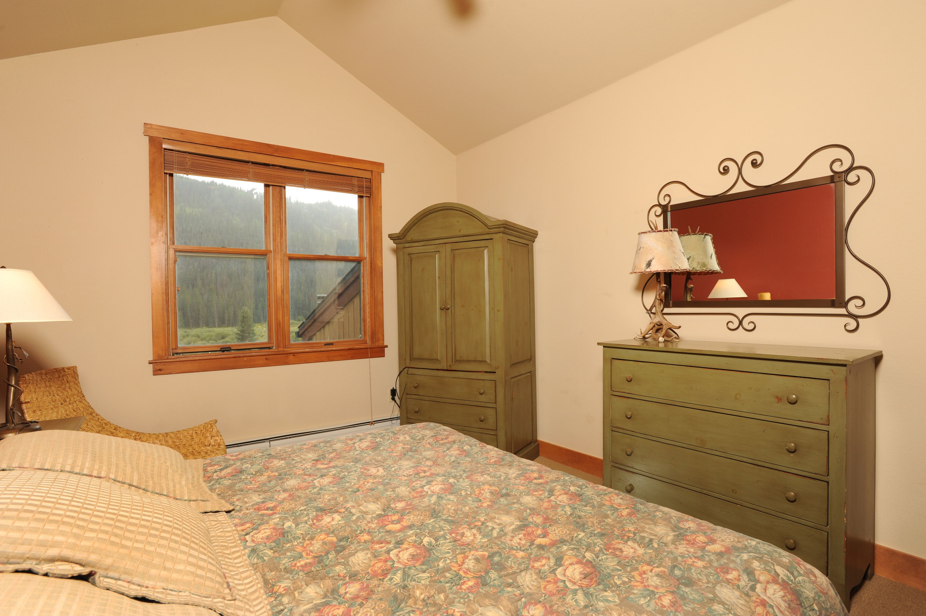 A bedroom with a bed, a dresser, a mirror, and a window with a view of a forest.