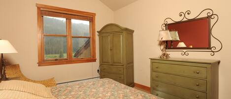 A bedroom with a bed, a dresser, a mirror, and a window with a view of a forest.