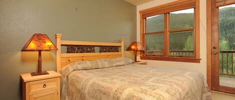 Quarto com cabeceira de madeira, cama com colcha estampada, duas luminárias de cabeceira, mesa de cabeceira com gaveta e janela com vista para uma floresta.