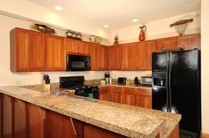 A kitchen with granite countertops, wooden cabinets, a black refrigerator, a microwave, a stove, and various kitchen appliances.