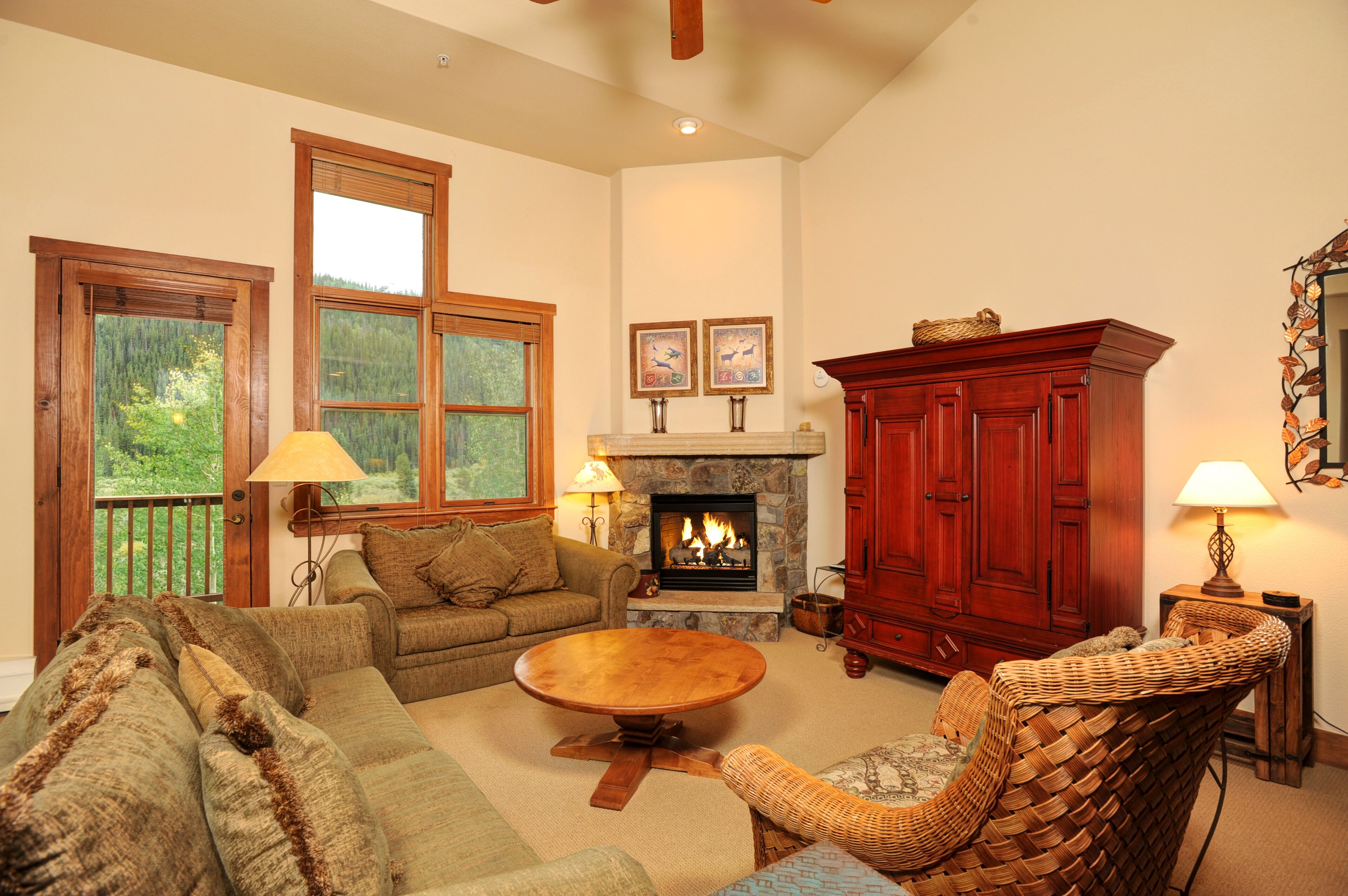 A cozy living room with a fireplace, wooden furniture, and large windows overlooking a forest.