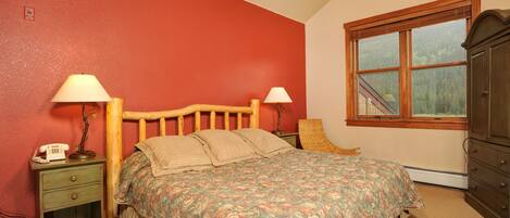 A bedroom with a wooden bed, two bedside tables, a nightstand, a window with a view of trees, and a red wall.