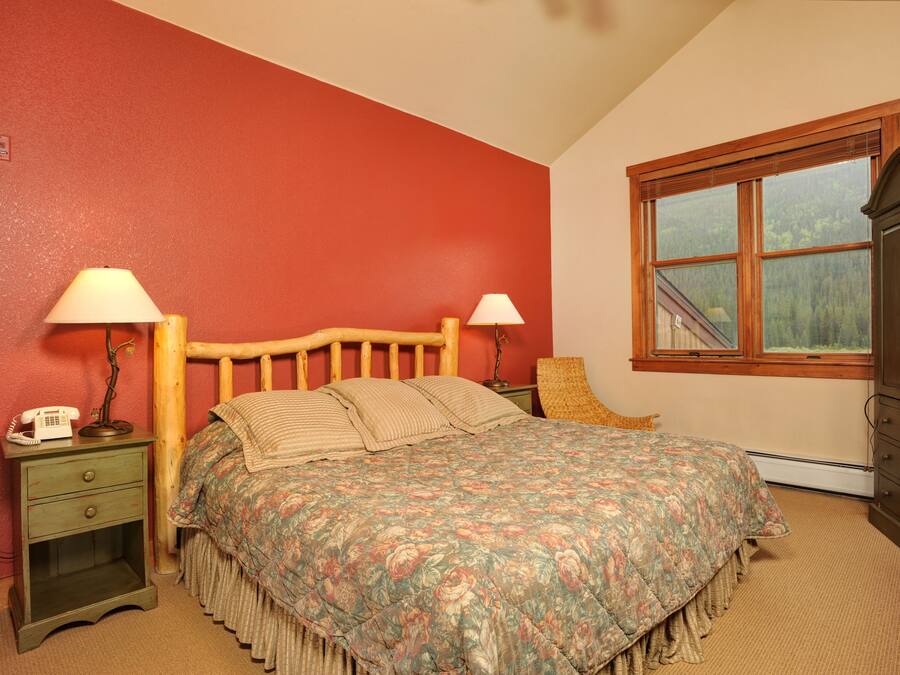 A bedroom with a wooden bed, two bedside tables, a nightstand, a window with a view of trees, and a red wall.