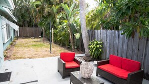 A patio area with wicker furniture and red cushions, surrounded by a wooden fence and tropical plants.