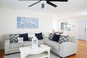 A modern living room with a grey sectional sofa, white coffee table, and a ceiling fan. A painting of dandelions hangs on the wall, and a small table with a lamp is in the background.